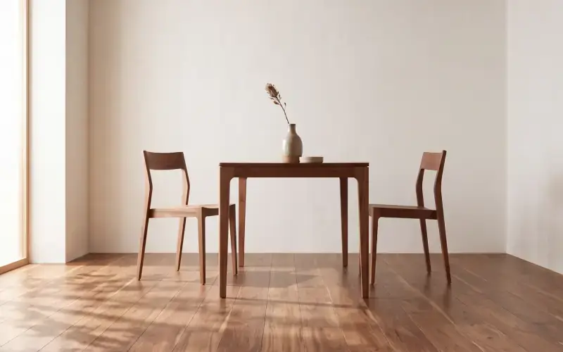 Small wooden dining table and two chairs on a warm brown plank floor against a plain cream wall