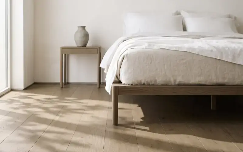Bright bedroom with light oak plank flooring, a linen-dressed wood bed and a pale vase on the nightstand