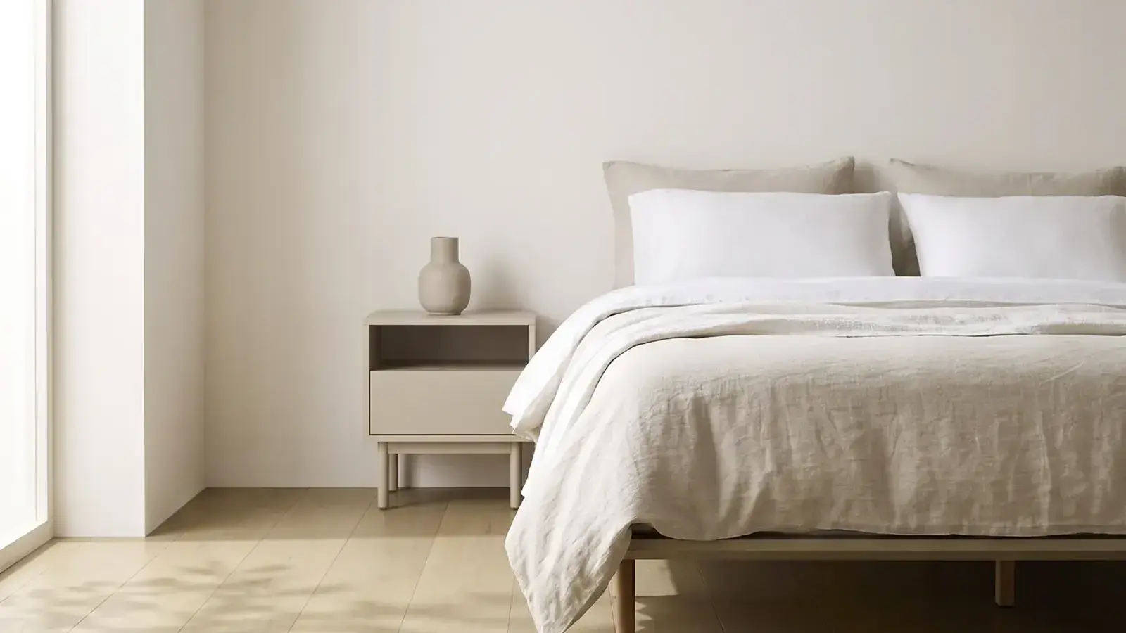 Bedroom with pale oak plank flooring, a linen covered bed and a beige nightstand beside a bright window