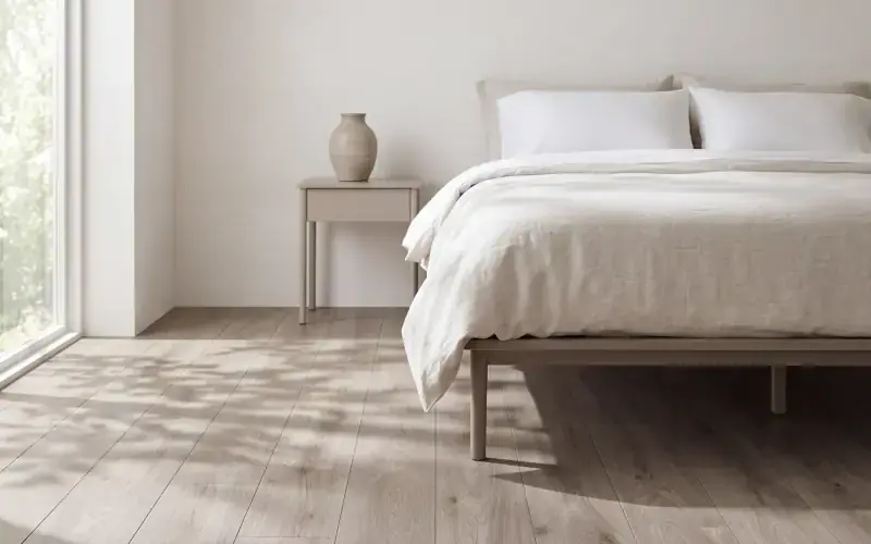 Pale taupe oak plank flooring in a bedroom with a linen bed and dappled light from a tall window
