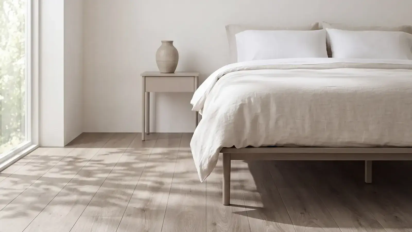 Pale taupe oak plank flooring in a bedroom with a linen bed and dappled light from a tall window