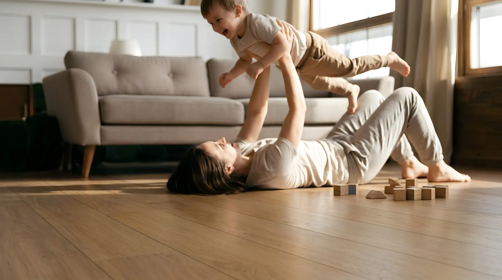 A parent lying on a warm oak floor, lifting a laughing child into the air